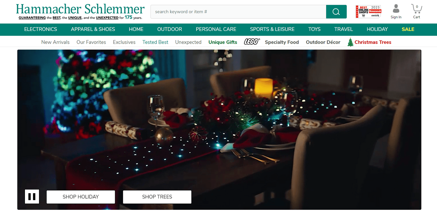 Festive dining table setup with holiday decorations and a glowing Christmas tree in the background at Hammacher Schlemmer.