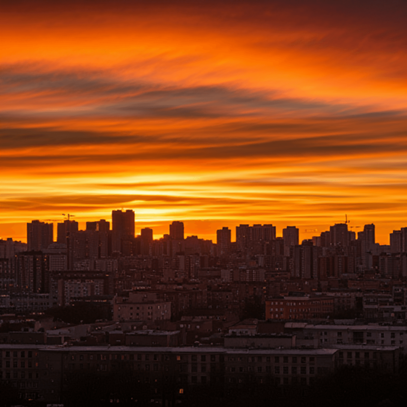 A vibrant orange sunset with dramatic clouds over a bustling city skyline silhouette.