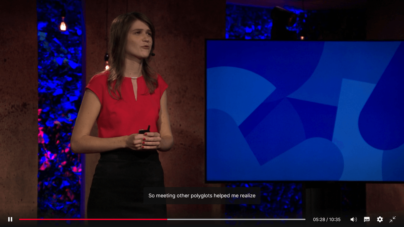 A female speaker in red top presenting a TED Talk with subtitles "So meeting other polyglots helped me realize.
