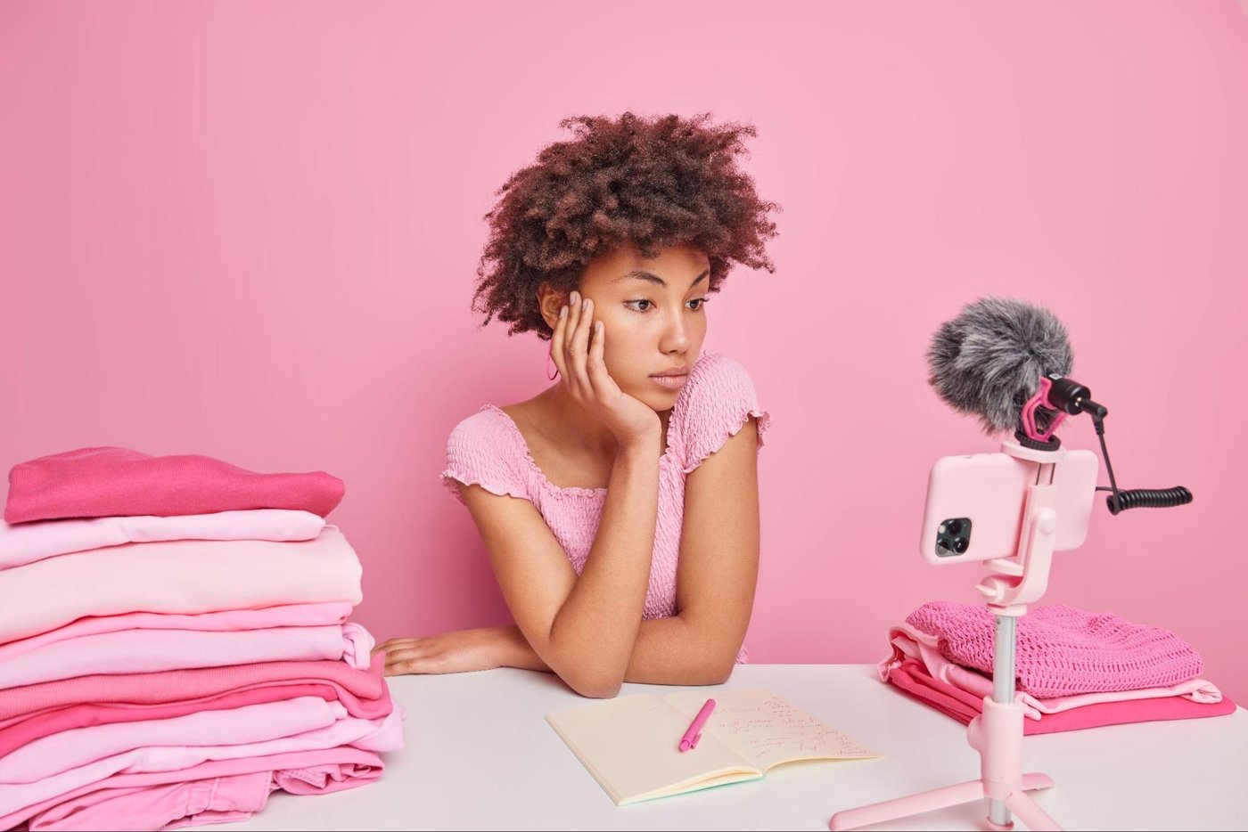 woman in pink shirt recording video with smartphone and microphone surrounded by folded clothes