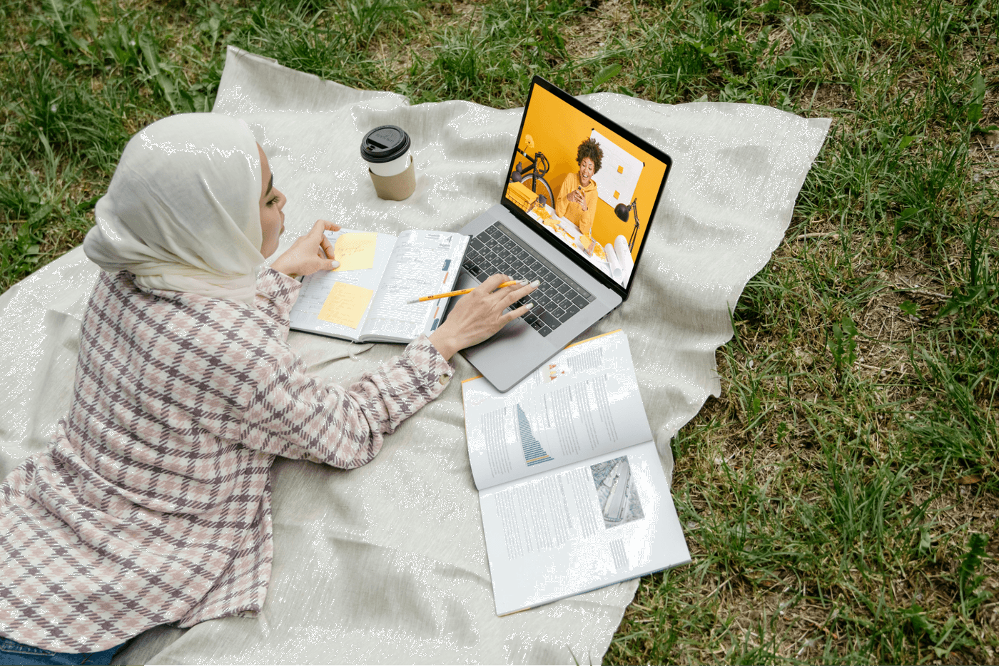 Woman in hijab studying outside on a blanket with laptop, notebook, and documents.