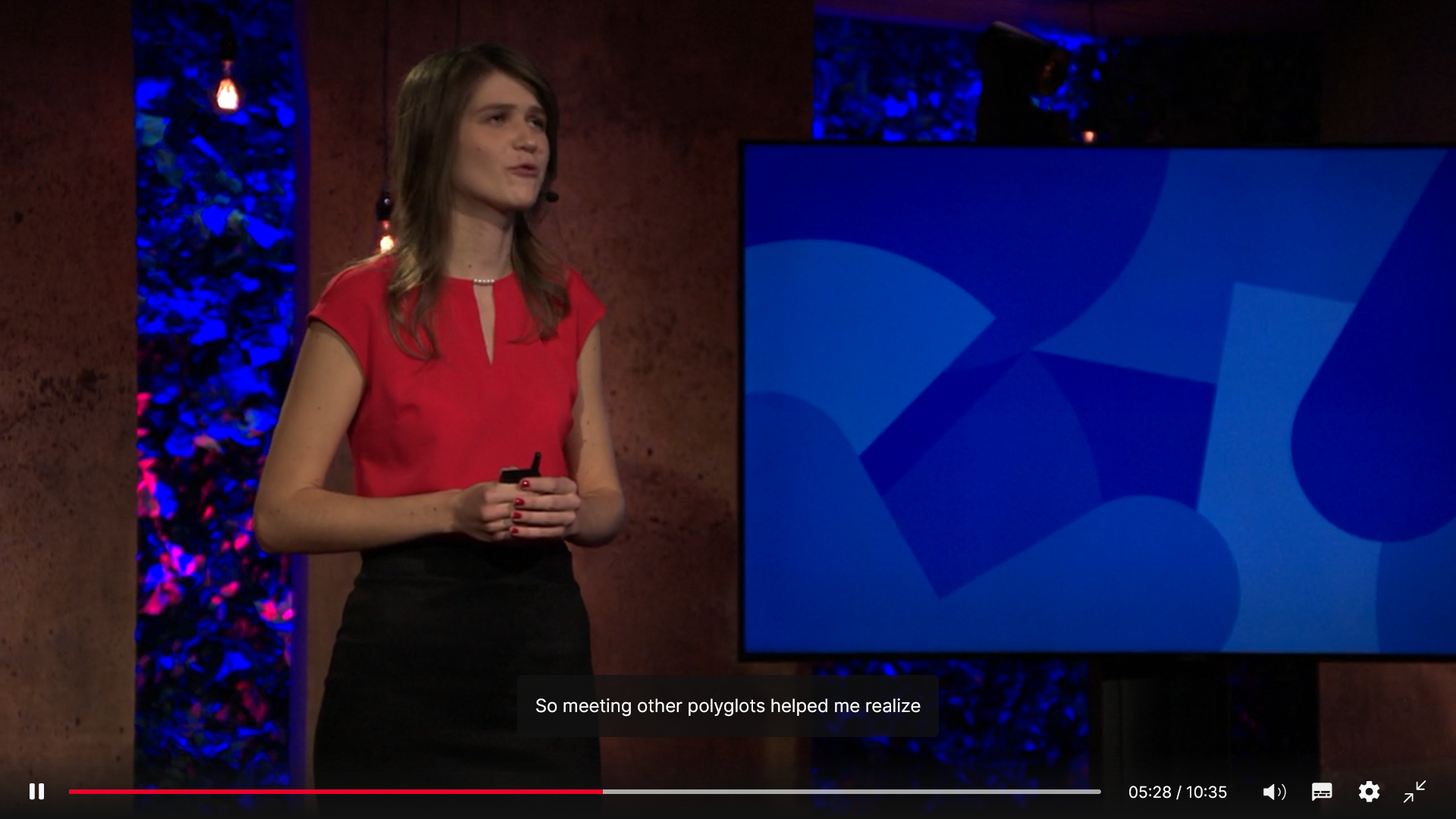 A female speaker in red top presenting a TED Talk with subtitles "So meeting other polyglots helped me realize.